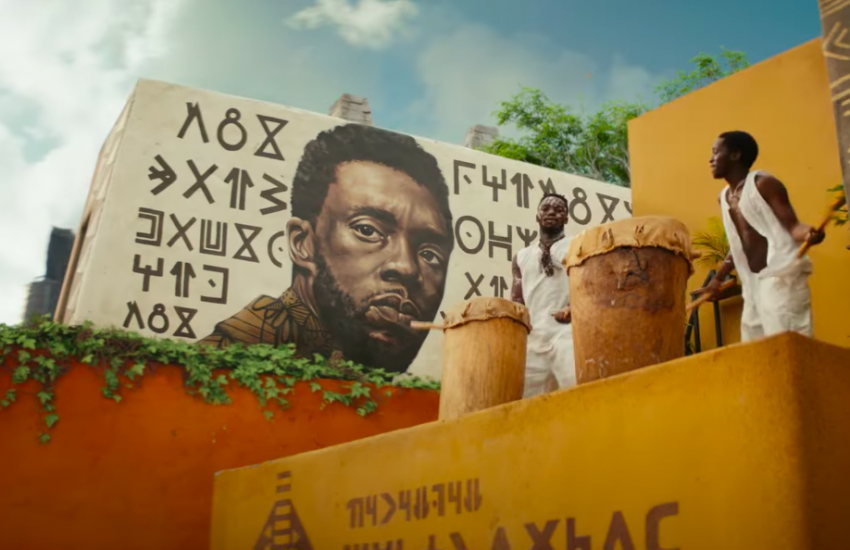 Movie still of two Wakandans playing drums in front of a mural of T'chala